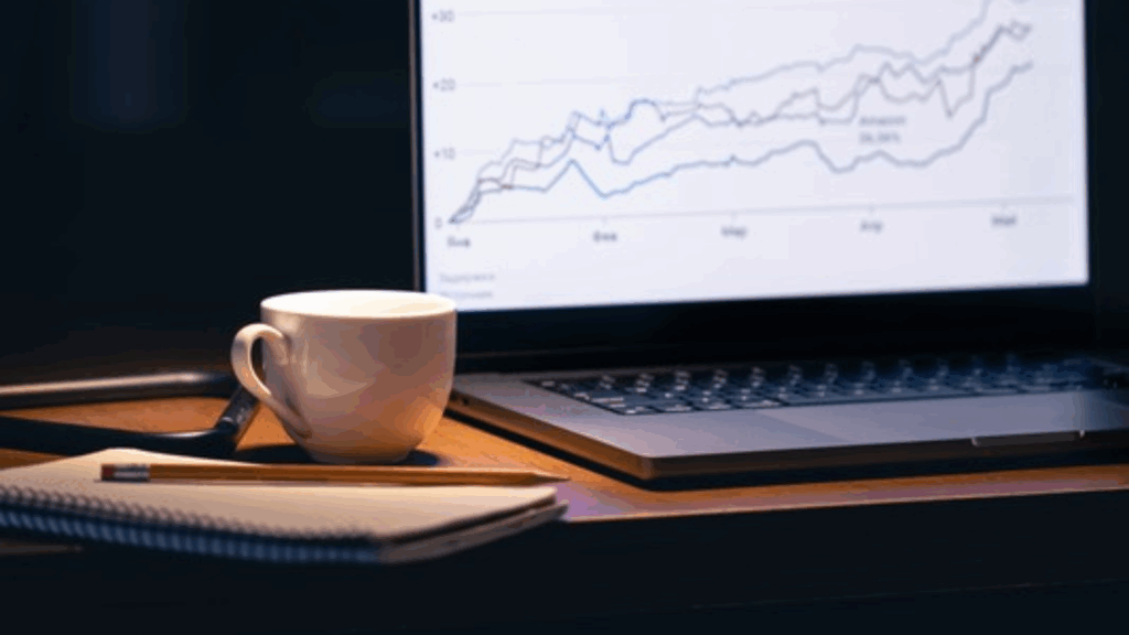 Laptop displaying line graph with rising trends on screen, desk with coffee cup and notebook in dimly lit workspace.
